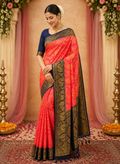 A woman models a traditional Indian saree in a decorated indoor setting. The saree has a vibrant coral-pink body with a navy blue border featuring intricate gold zari embroidery, paired with a solid navy blue blouse.