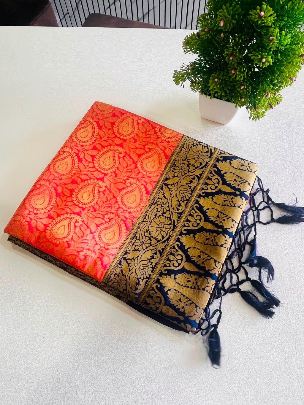 A folded red and gold brocade scarf with a navy blue and gold patterned border and black tassels, displayed on a white surface next to a small green potted plant.