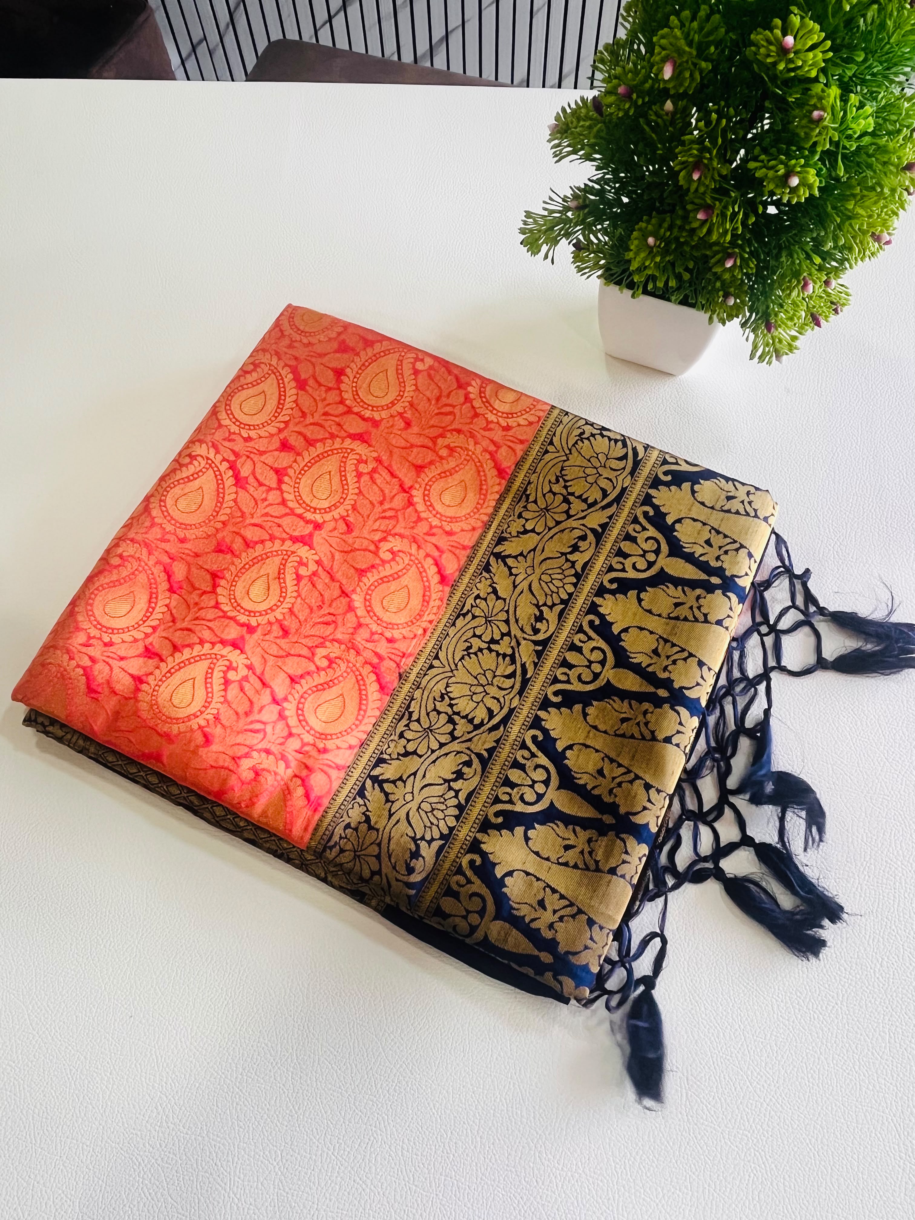 A folded red and gold brocade scarf with a navy blue and gold patterned border and black tassels, displayed on a white surface next to a small green potted plant.
